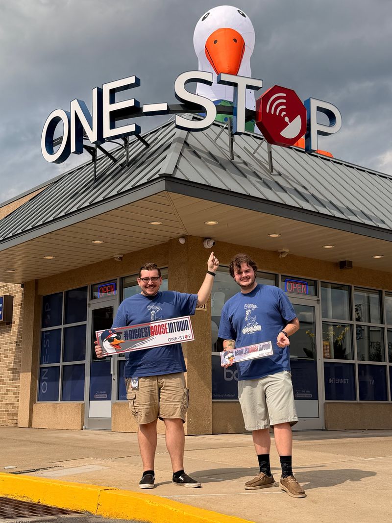 Two One-Stop team members holding #BiggestGooseInTown and #SmallestGooseInTown signs in front of the One-Stop office, with the giant goose on the roof