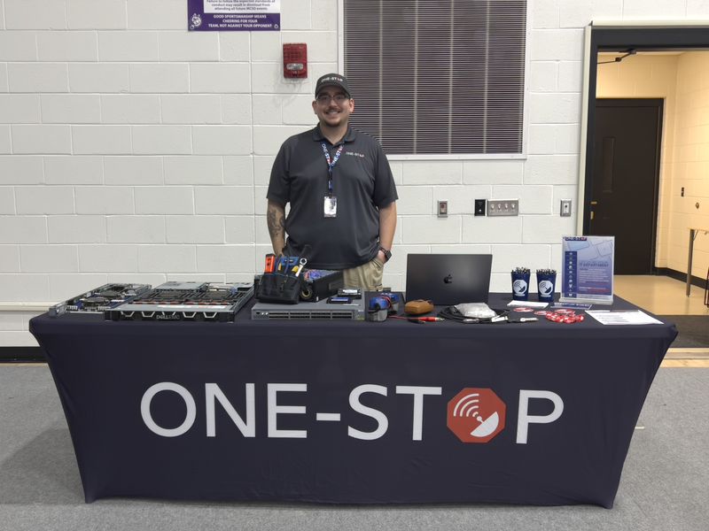 A One-Stop team member standing behind the One-Stop table at Mifflin County High School Career Day, with servers, a network switch and tools on display