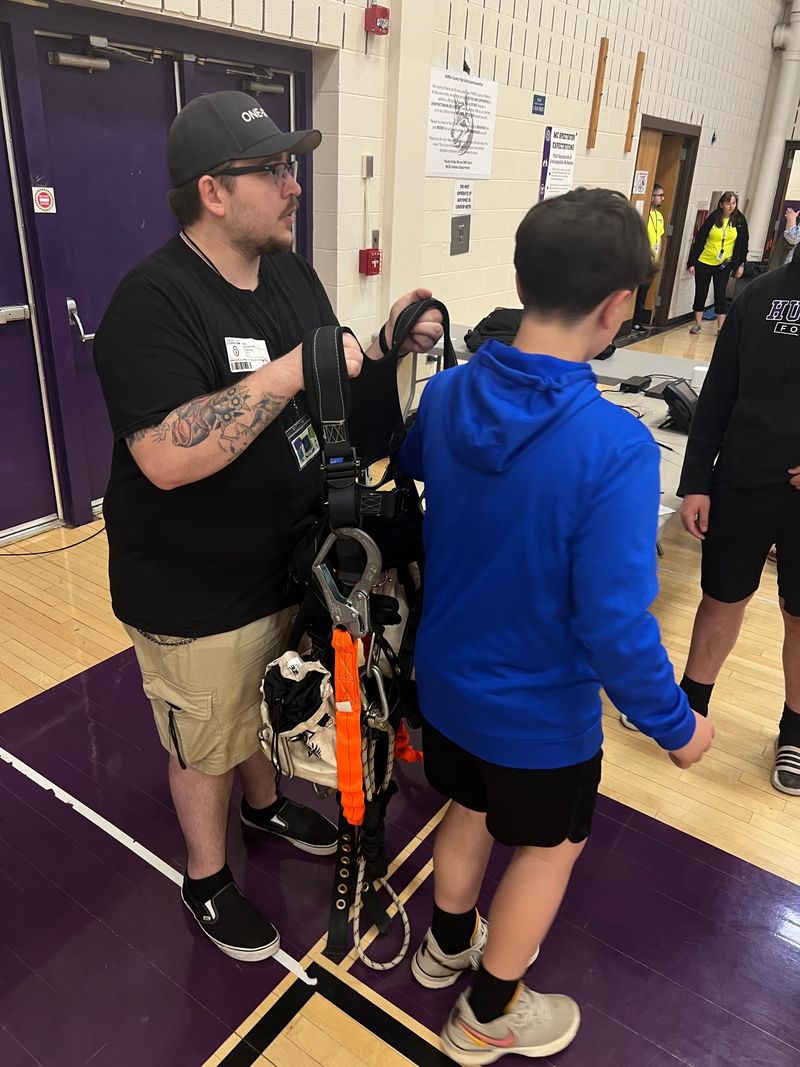 A One-Stop technician holding up a tower climbing harness for a student at Career Day