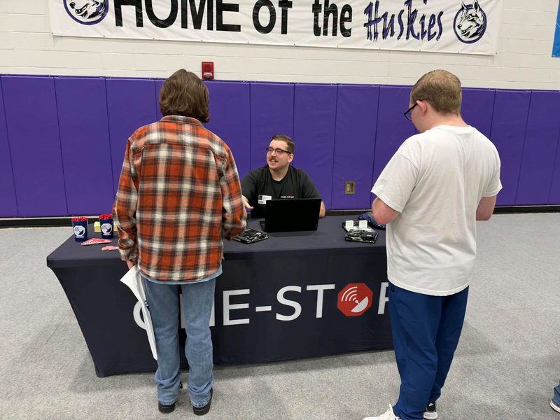 A One-Stop team member at the One-Stop table talking with two students at Mifflin County High School Career Day, with computer parts and a laptop on display in the gym