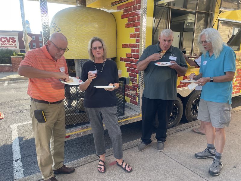 Chamber guests eating pizza beside a yellow wood-fired pizza food truck outside the One-Stop office