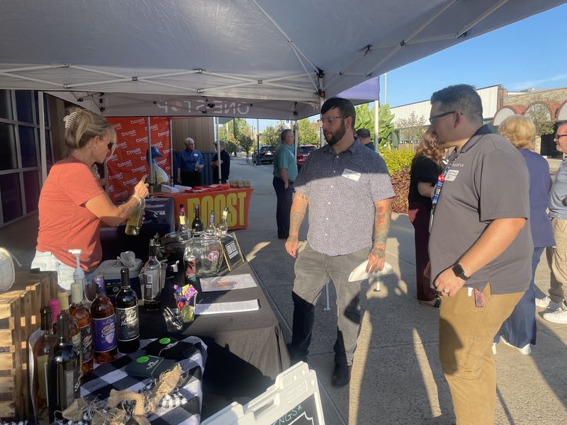 Guests visiting a local vendor table under the One-Stop tent at the Chamber After Hours event, with the Boost Mobile booth behind