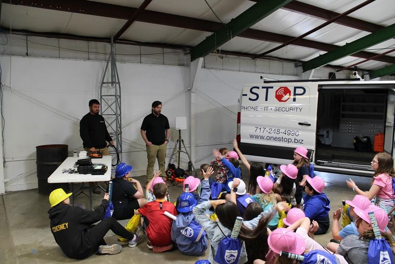 Two One-Stop technicians talking with a group of students in pink, blue and yellow hard hats next to a One-Stop van at Free Wheeling Day