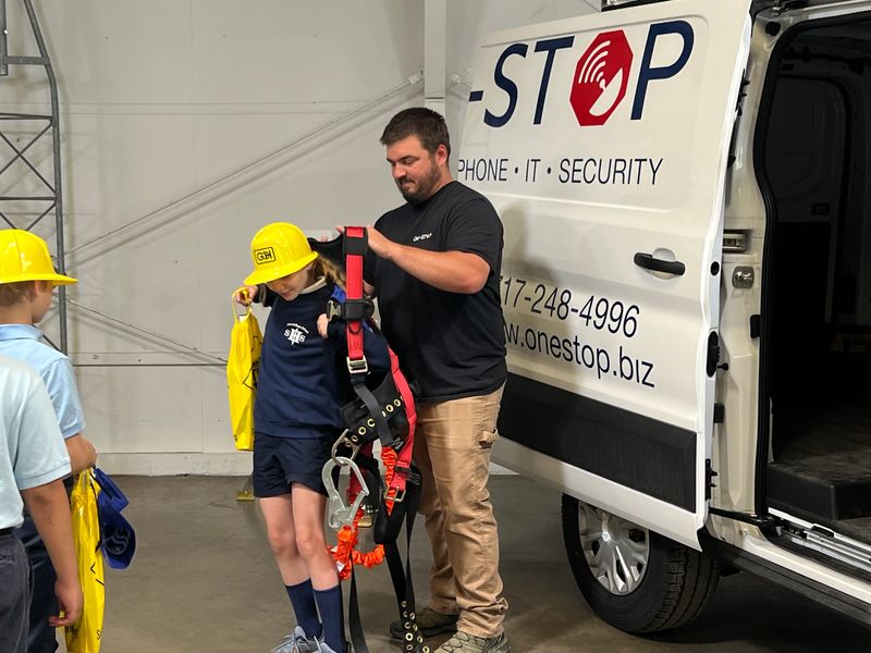 A One-Stop technician helping a student in a yellow hard hat try on a tower climbing harness beside a One-Stop van