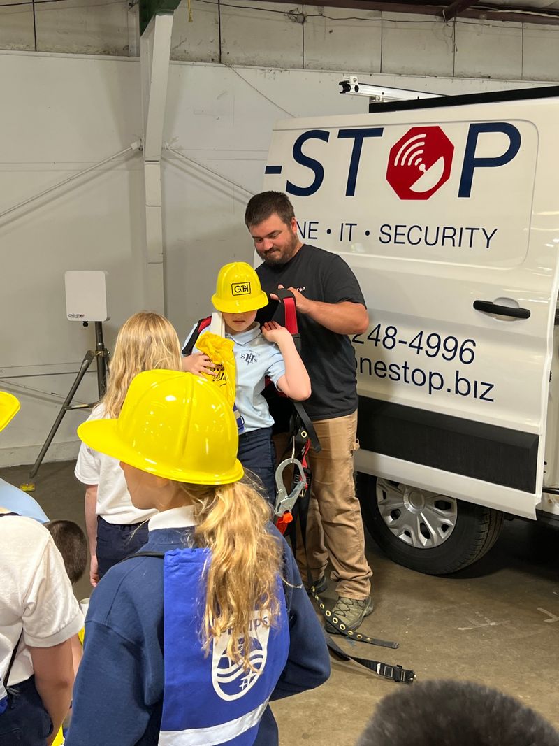 Students in yellow hard hats waiting while a One-Stop technician fits a climbing harness on a classmate