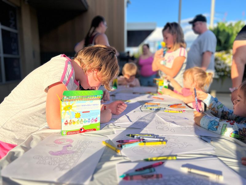Children coloring Goose Day and One-Stop coloring pages with crayons at a long table outside the One-Stop office during Lucky Friday