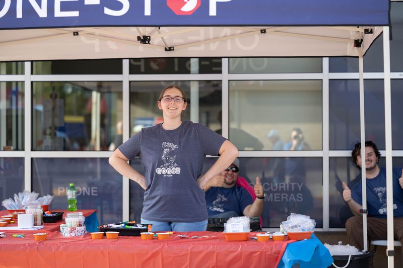 A One-Stop team member smiling behind the kids’ craft table under the One-Stop tent at Lucky Friday, with two coworkers giving a thumbs up