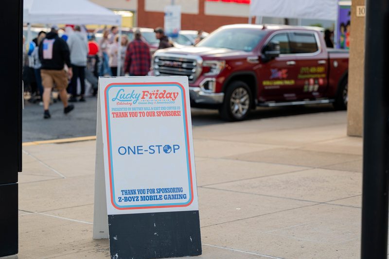 Lucky Friday 2026 sponsor sign thanking One-Stop on the sidewalk in downtown Lewistown, with the event crowd behind it