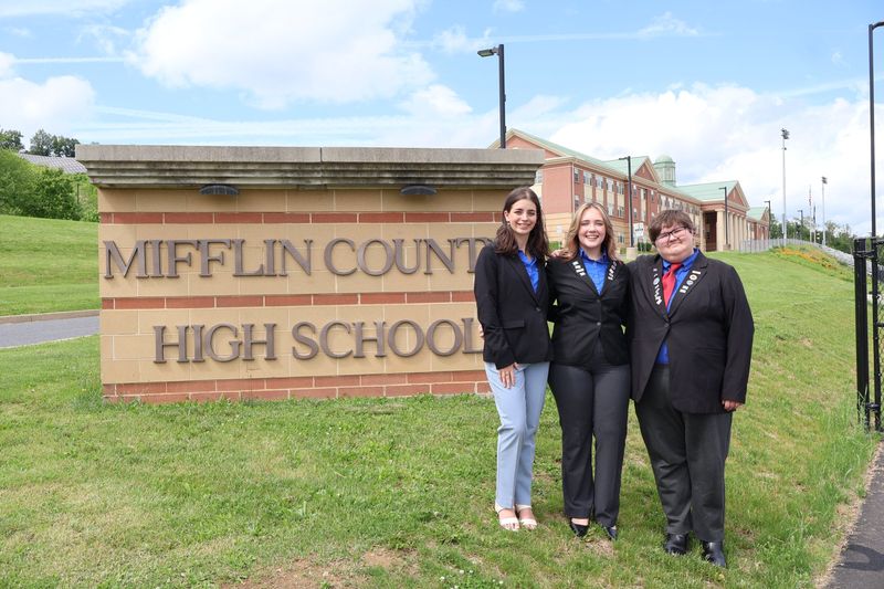 Three Mifflin County High School Technology Student Association members in blazers standing beside the Mifflin County High School sign