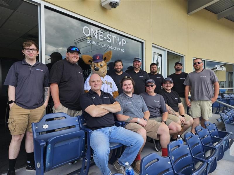 The One-Stop team posing with the State College Spikes mascot in front of the One-Stop Communications suite at Medlar Field